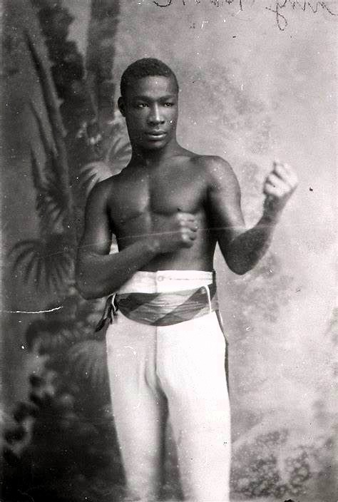 A historical photograph of a male boxer, showcasing a strong physique with bare torso and fighting stance, wearing light trousers and a sash around the waist, set against a backdrop of palm leaves.