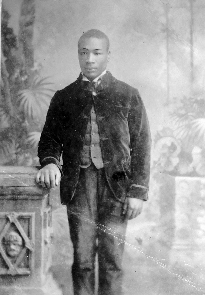 A vintage black and white photograph of a young man in formal attire, standing beside an ornate pedestal. The background features foliage and decorative elements, suggesting a studio setting. The subject appears composed and serious.