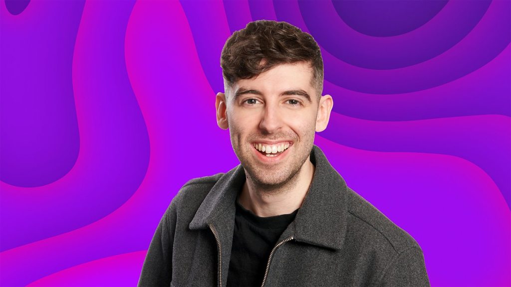 A smiling young man with short hair, wearing a black shirt and a gray jacket, against a vibrant purple and abstract background.