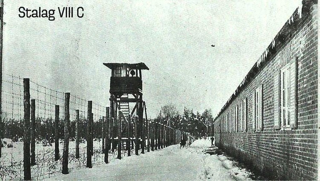 Black and white photograph of Stalag VIII C, featuring a watchtower and bordering barbed wire fencing in a snowy landscape.