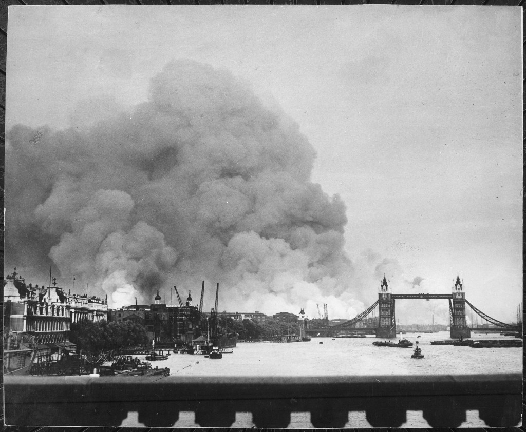 A historic black and white photograph showing thick smoke rising from buildings in the East End of London, with the iconic Tower Bridge in the foreground and boats in the river Thames.